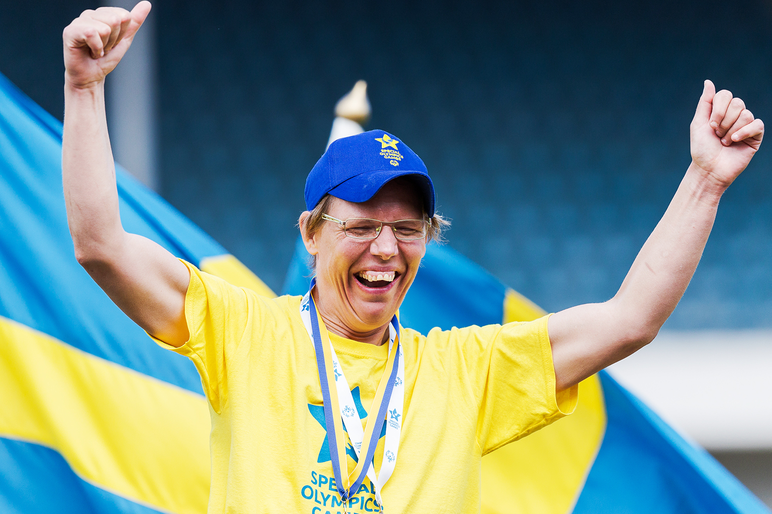 Foto, utomhus på friidrottsarena. En person håller upp armarna i segergest, medalj runt halsen, Sverigeflaggor och läktare bakom.