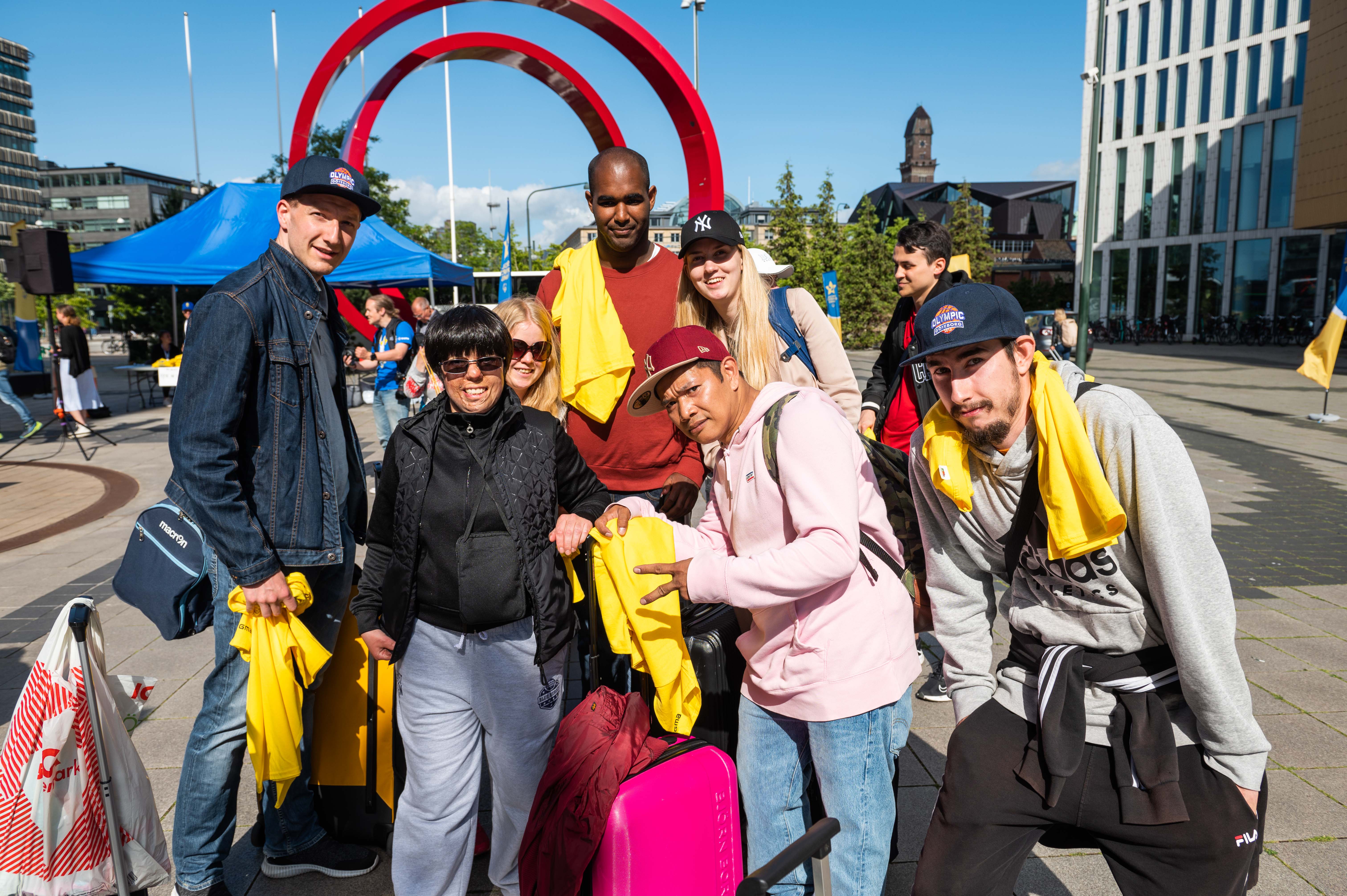 Foto, utomhus. En grupp står på ett torg framför en röd båge.